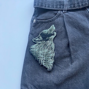 May include: A pair of dark gray denim jeans with a green embroidered wolf patch on the front pocket.