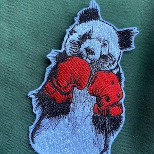 Puede incluir: Parche bordado de un panda con guantes de boxeo rojos. El panda es blanco con marcas negras y tiene una expresión seria.