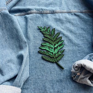 May include: A light blue denim jacket featuring a green and black embroidered fern patch. The fern patch is intricately detailed with shades of green and black thread. The sleeves are rolled up, revealing a grey lining.
