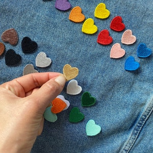 May include: A variety of colorful felt heart patches arranged on a blue denim background. The patches are in different colors, including red, blue, green, yellow, orange, purple, and pink. A hand is holding a gold heart patch.