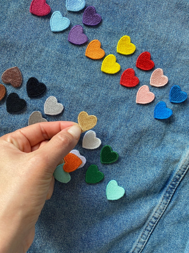May include: A variety of colorful felt heart patches arranged on a blue denim background. The patches are in different colors, including red, blue, green, yellow, orange, purple, and pink. A hand is holding a gold heart patch.