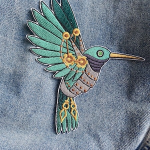 Puede incluir: Un parche bordado que representa un colibrí en vuelo, con intrincados detalles en verde y dorado. El diseño tiene una estética steampunk.