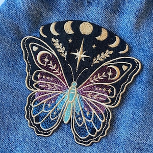 May include: Iron-on patch featuring an embroidered black butterfly with gold and purple accents. The butterfly has a crescent moon and star design on its wings.