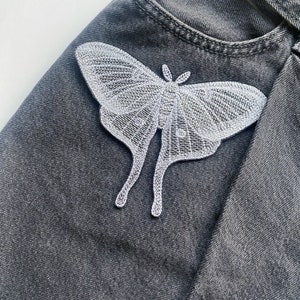 May include: A white embroidered moth applique on a dark gray denim fabric.