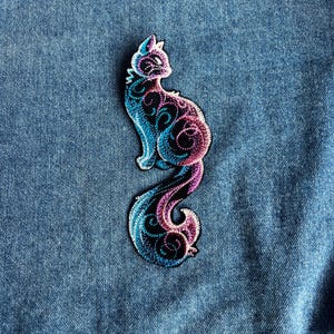 Puede incluir: Parche bordado de gato en tonos azules, morados y negros. El gato está representado en una pose sentada con una larga cola fluida. El diseño presenta intrincados patrones arremolinados.