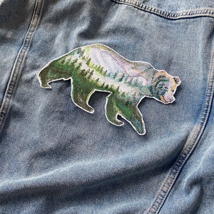 May include: An embroidered patch of a bear with a mountain landscape inside its silhouette. The patch is green, brown and white and is sewn onto a blue denim jacket.