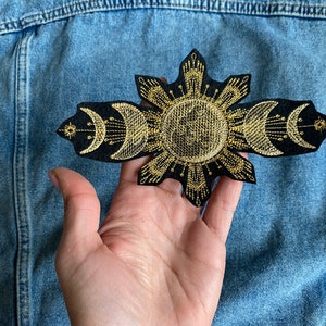 Op de afbeelding: Een zwarte geborduurde patch met een gouden zon- en maanontwerp. De zon is rond met stralen die er vanaf komen. De maan is een halve maan en wordt drie keer herhaald.
