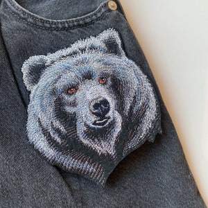 Peut inclure: Un gros plan d'un écusson de ours brodé gris sur une veste en jean noire. La fourrure de l'ours est détaillée avec des points blancs, et ses yeux sont bruns.