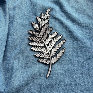 May include: Embroidered fern patch in shades of gray and white, with a black border, applied to a light blue denim fabric. The fern design is detailed and textured, creating a naturalistic aesthetic. Ideal for adding a botanical touch to clothing or accessories.