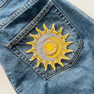Può includere: Toppa ricamata con un design celeste di sole e luna su una tasca di jeans. Il sole è giallo con raggi, e la luna è argentata con dettagli intricati. La toppa è cucita su tessuto denim blu.
