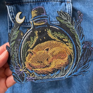 May include: Embroidered patch featuring a sleeping fox inside a bottle, surrounded by a crescent moon and foliage. The patch has a black background with colorful stitching in shades of gold, blue, green, and purple. The patch is on a denim jacket.