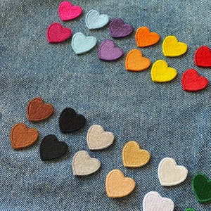 May include: Assortment of felt heart patches in various colors, including red, pink, blue, purple, orange, yellow, brown, black, gray, and green. The patches are arranged in a semi-circle on a blue denim background.