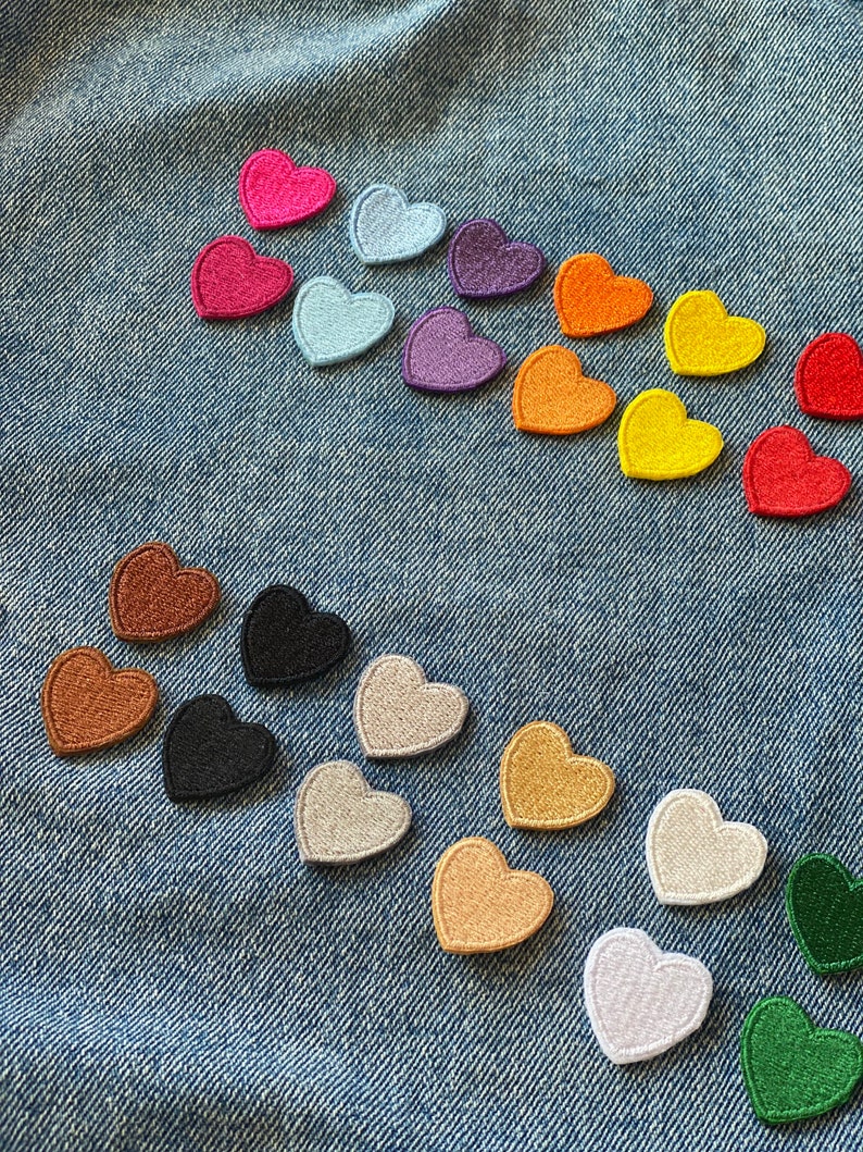 May include: Assortment of felt heart patches in various colors, including red, pink, blue, purple, orange, yellow, brown, black, gray, and green. The patches are arranged in a semi-circle on a blue denim background.