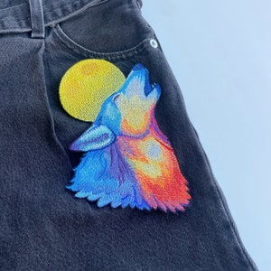 Puede incluir: Un lobo bordado de color azul y naranja aullando a la luna sobre un fondo de mezclilla negra.