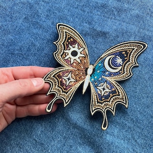 Puede incluir: Un parche de mariposa bordado en dorado y azul con un diseño de sol, luna y estrellas.