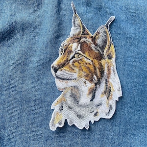 May include: A close-up of an embroidered patch featuring a brown and white lynx with large ears. The patch is on a blue denim background.