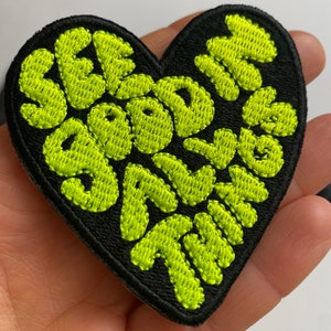 Op de afbeelding: Een zwarte hartvormige patch met de woorden "See Good in All Things" geborduurd in neon groen.