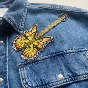 Könnte beinhalten: Ein gelber und orangefarbener Schmetterlingsgitarren-Patch auf einer blauen Jeansjacke.