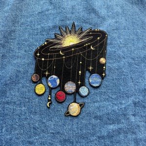 May include: Embroidered patch with a celestial design, including a sun, planets, and stars. The patch is black with gold and colourful accents, including yellow, blue, red, and brown. The patch is likely for clothing or accessories.