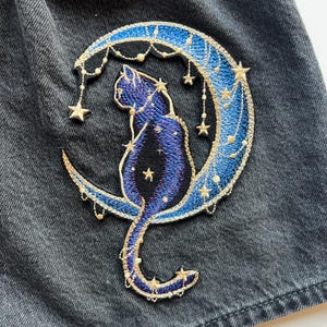 Könnte beinhalten: Gestickter Aufnäher mit einer blauen Katzen-Silhouette auf einem Halbmond. Der Mond ist mit goldenen und blauen Fäden umrandet, mit hängenden goldenen Sternen. Katze und Mond vor dunklem, grauem Hintergrund.