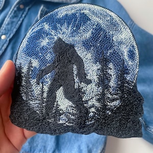 Könnte beinhalten: Gestickter Aufnäher mit einer schwarzen Silhouette von Bigfoot, der vor einem Vollmond geht. Der Mond ist blau und weiß mit einem strukturierten Design.