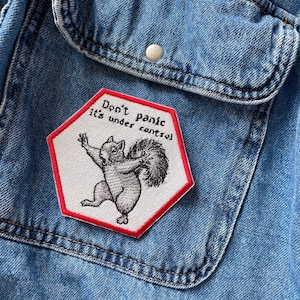 May include: A white embroidered patch with a red border in the shape of a hexagon. The patch features a cartoon squirrel with the text "Don't panic it's under control".