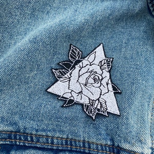 Puede incluir: Rosa blanca bordada con hojas en forma de triángulo sobre una chaqueta vaquera azul.