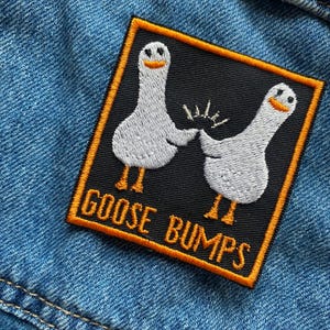Op de afbeelding: Een geborduurde patch in zwart en wit met twee witte ganzen die elkaars hand vasthouden. De patch heeft een oranje rand en de tekst "Goose Bumps" in oranje.