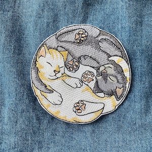 May include: Embroidered patch of two cats curled up together in a yin yang symbol. The cat on the left is white with orange markings and the cat on the right is black.