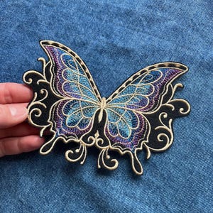 Puede incluir: Un parche bordado con una mariposa azul y morada con hilo dorado y tela negra. La mariposa tiene detalles intrincados y un diseño de remolino a su alrededor.