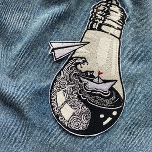 Può includere: Una toppa ricamata in bianco e nero che rappresenta una lampadina con una piccola barca a vela al suo interno. La lampadina è piena di onde e un aeroplano di carta esce dalla parte superiore.