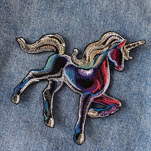 Peut inclure: Patch brodé thermocollant représentant une licorne noire, bleue et violette avec une crinière et une corne dorées. La licorne court la tête tournée sur le côté.