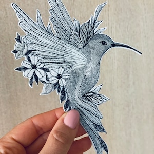 Puede incluir: Un parche bordado gris que representa un colibrí en vuelo con las alas extendidas. El colibrí está rodeado de detalles florales blancos, creando un diseño delicado e intrincado.