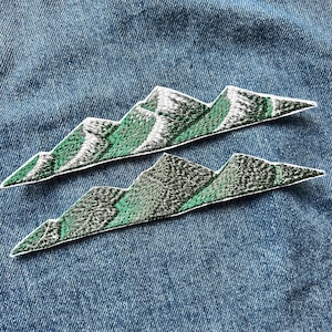 May include: Two embroidered patches featuring a mountain range design. Each patch displays a jagged, elongated silhouette with green slopes and white, textured peaks. The embroidery uses dense stitching to create depth and contrast between the green base and the white highlights on the mountain tops, finished with a clean white border.