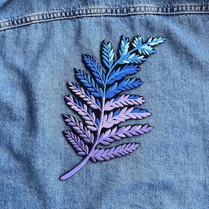 May include: A detailed embroidered leaf patch in shades of blue and purple, affixed to a denim jacket. The patch features a gradient effect, transitioning from light blue to deep purple. The leaf design is intricate and detailed.