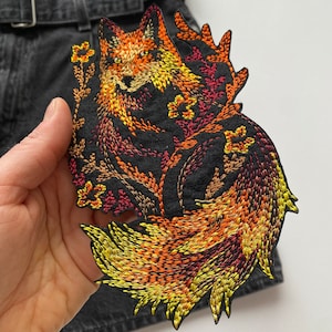 Può includere: Toppa ricamata con una volpe nei toni dell'arancione, giallo e rosso su sfondo nero. La volpe è circondata da elementi floreali. La toppa è probabilmente per abbigliamento o accessori.