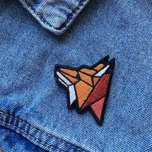 May include: A geometric fox-shaped embroidered patch in orange, red, and white colors, sewn onto a blue denim jacket.