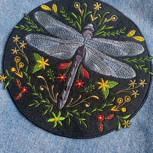 Peut inclure: Un patch en feutre noir avec une libellule brodée entourée de fleurs colorées. La libellule est bleue et violette avec des fleurs vertes et jaunes.