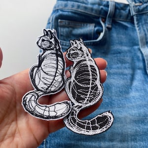 Puede incluir: Dos parches de hierro bordados en blanco y negro de gatos. Los gatos están en posición sentada con sus colas enroscadas alrededor de sus cuerpos.