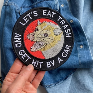 Può includere: Una toppa ricamata in bianco e nero con sfondo rosso. La toppa presenta un opossum dei cartoni animati con il testo "Let's Eat Trash And Get Hit By A Car".
