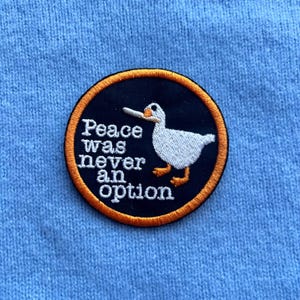 Op de afbeelding: Geborduurde patch met een zwarte achtergrond en een oranje rand. De patch toont een witte cartoon eend en de tekst "Peace was never an option" in het wit. De patch zit op een lichtblauwe stof.