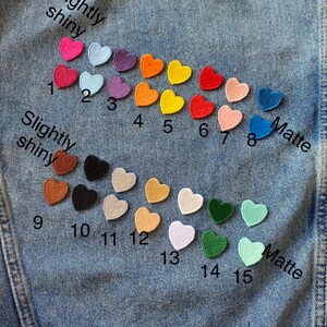 May include: Fifteen felt heart patches in various colors, including red, pink, orange, yellow, green, blue, and brown. The patches are arranged in two rows on a blue denim background. The top row is labeled "Slightly shiny" and the bottom row is labeled "Matte".