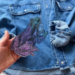 May include: A blue denim jacket with a colorful embroidered frog patch on the front. The frog is iridescent and has a black outline.