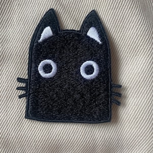 May include: Black embroidered iron-on patch of a cat with white eyes and white markings on its ears.