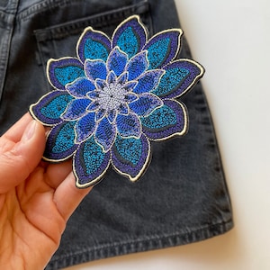 Könnte beinhalten: Ein gestickter Blumen-Patch in Blau und Lila mit goldener Umrandung. Der Patch befindet sich auf einem dunkelblauen Jeansstoff.