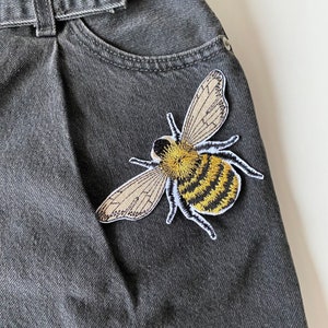 Op de afbeelding: Een zwart denim jack met een geborduurde bij-patch in geel en bruin op de zak.