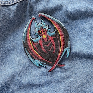 Puede incluir: Parche bordado con un dragón estilizado con cuerpo negro, alas rojas y detalles azules y amarillos. El dragón tiene un diseño detallado con cuernos y una expresión feroz. El parche está adherido a una tela vaquera azul.
