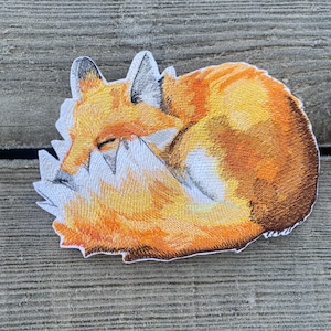 Puede incluir: Un parche bordado que representa un zorro naranja enroscado con acentos blancos. El zorro está durmiendo con los ojos cerrados.