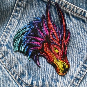 Może przedstawiać: Kolorowa haftowana naszywka z głową smoka z tęczowymi łuskami i czarnym tłem. Naszywka jest przyszyta do niebieskiej kieszeni dżinsowej.