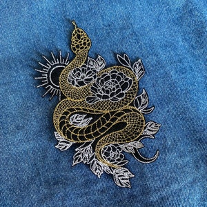 Puede incluir: Un parche bordado en oro y negro con una serpiente enroscada alrededor de una flor con un sol detrás.